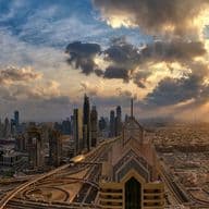 A panoramic view of Dubai at sunset, featuring skyscrapers, including a prominent tall tower, under a dramatic sky with sun rays.