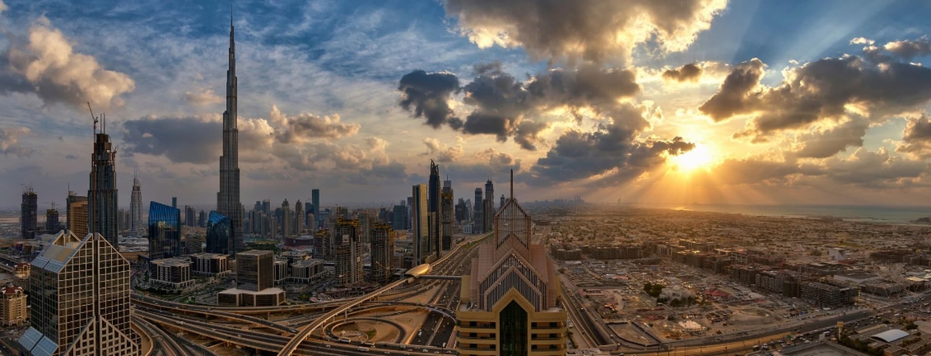 A panoramic view of Dubai at sunset, featuring skyscrapers, including a prominent tall tower, under a dramatic sky with sun rays.