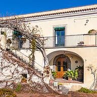 Eine Villa im mediterranen Stil mit cremefarbener Fassade, Rundbögen, Balkonen und Kletterpflanzen unter einem strahlend blauen Himmel.