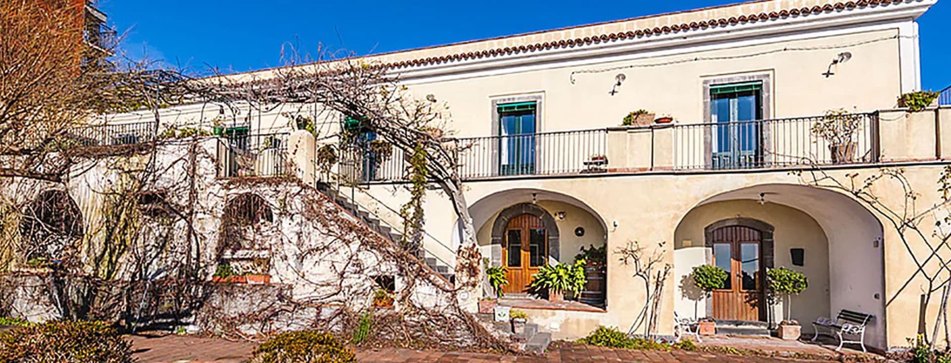 Eine Villa im mediterranen Stil mit cremefarbener Fassade, Rundbögen, Balkonen und Kletterpflanzen unter einem strahlend blauen Himmel.