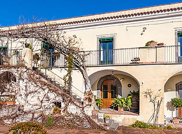 A Mediterranean-style villa with a cream facade, arched doorways, balconies, and climbing plants under a clear blue sky.