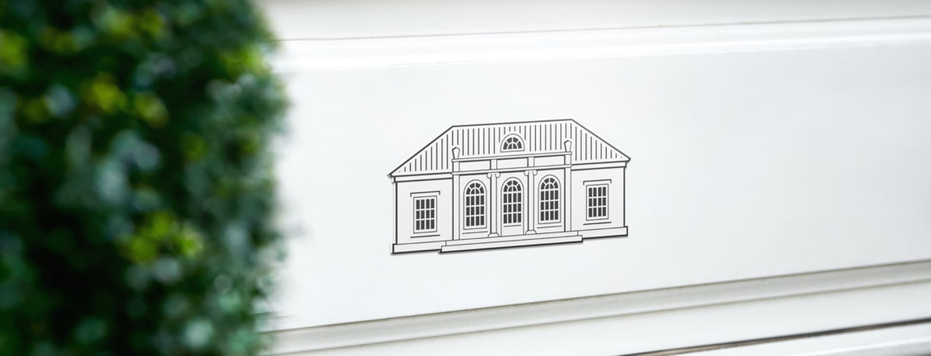 Engel & Völkers Villa Logo auf einer weißen Wand. Auf der linken Seite ist leicht verschwommen ein grüner Buchsbaum.