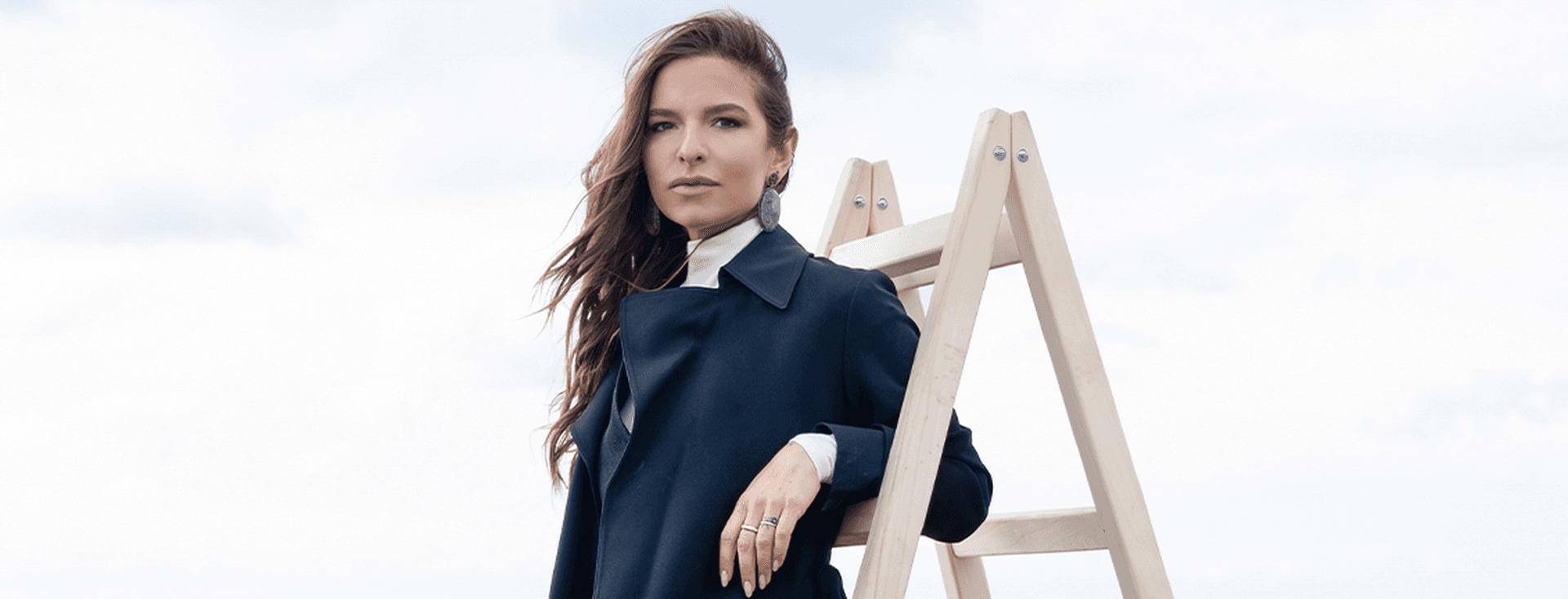 Γυναίκα με μαύρο κομψό παλτό ποζάρει δίπλα σε ξύλινη σκάλα σε μινιμαλιστικό θαλάσσιο σκηνικό.