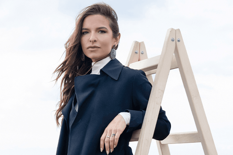Γυναίκα με μαύρο κομψό παλτό ποζάρει δίπλα σε ξύλινη σκάλα σε μινιμαλιστικό θαλάσσιο σκηνικό.