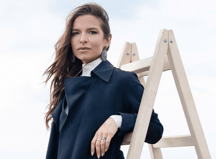 Γυναίκα με μαύρο κομψό παλτό ποζάρει δίπλα σε ξύλινη σκάλα σε μινιμαλιστικό θαλάσσιο σκηνικό.