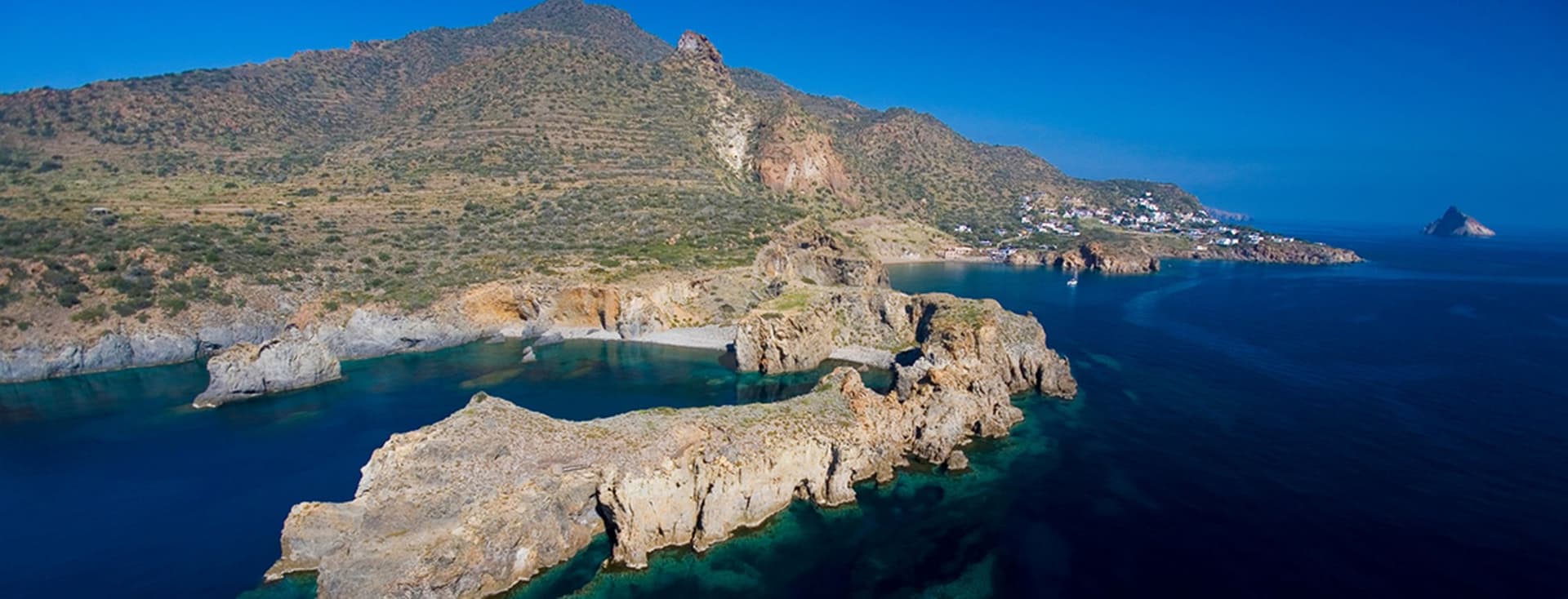 Luftaufnahme einer zerklüfteten Küste mit felsigen Klippen, klarem blauem Wasser und einer entfernten Küstenstadt unter klarem Himmel.