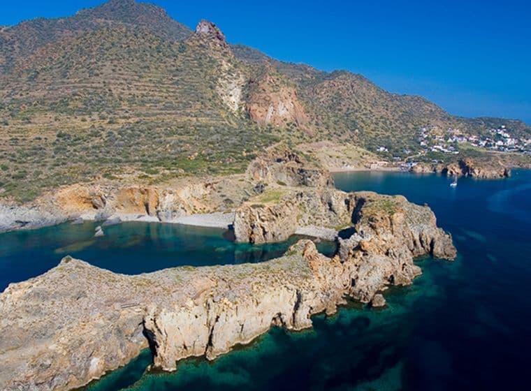 Aerial view of a rugged coastline with rocky cliffs, clear blue waters, and a distant coastal town under a clear sky.
