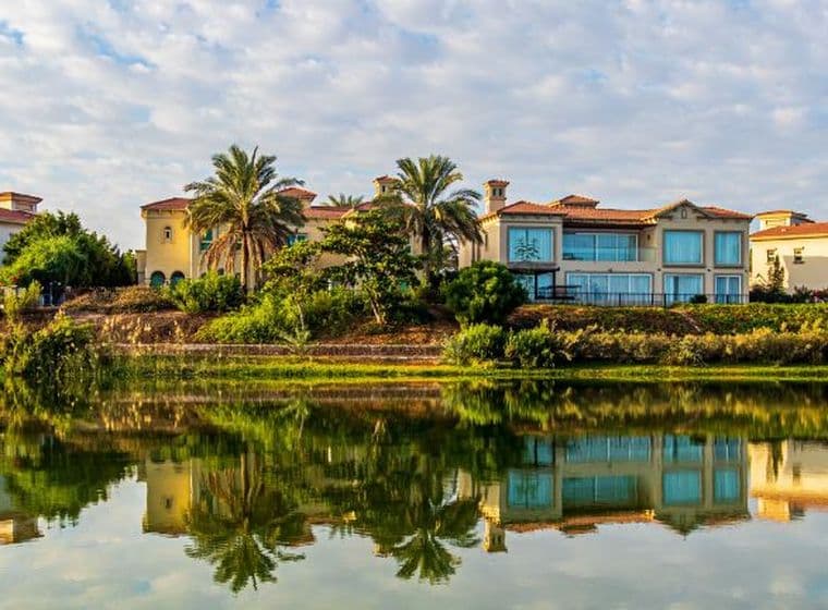 Row of luxurious villas with palm trees by a calm lake, reflecting in the water under a partly cloudy sky.