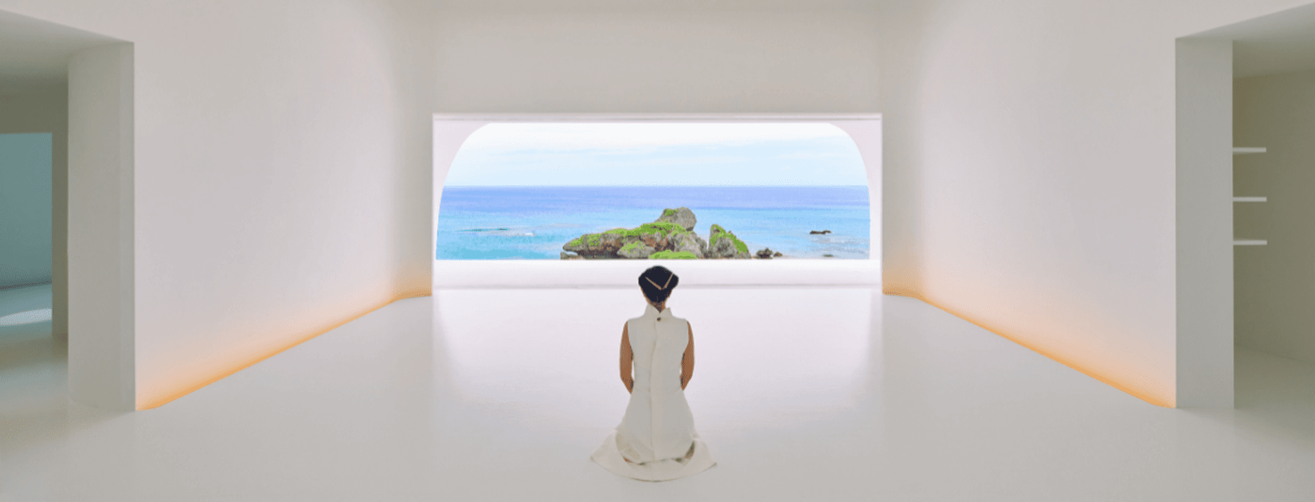 Eine Person in einem weißen Kleid kniet in einem minimalistisch gestalteten weißen Raum und blickt durch ein großes Bogenfenster auf das Meer.
