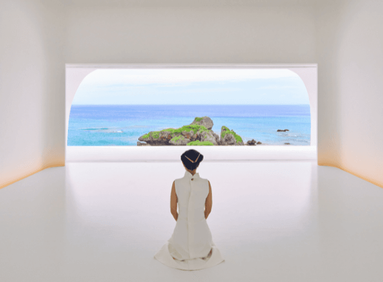 A person in a white dress kneels in a minimalist white room, facing a sea view through a large arched window.