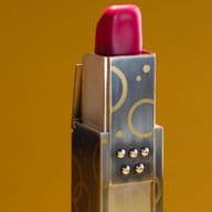 Roter Lippenstift mit Etui aus Gold und Silber mit geometrischem Muster vor goldbraunem Hintergrund.