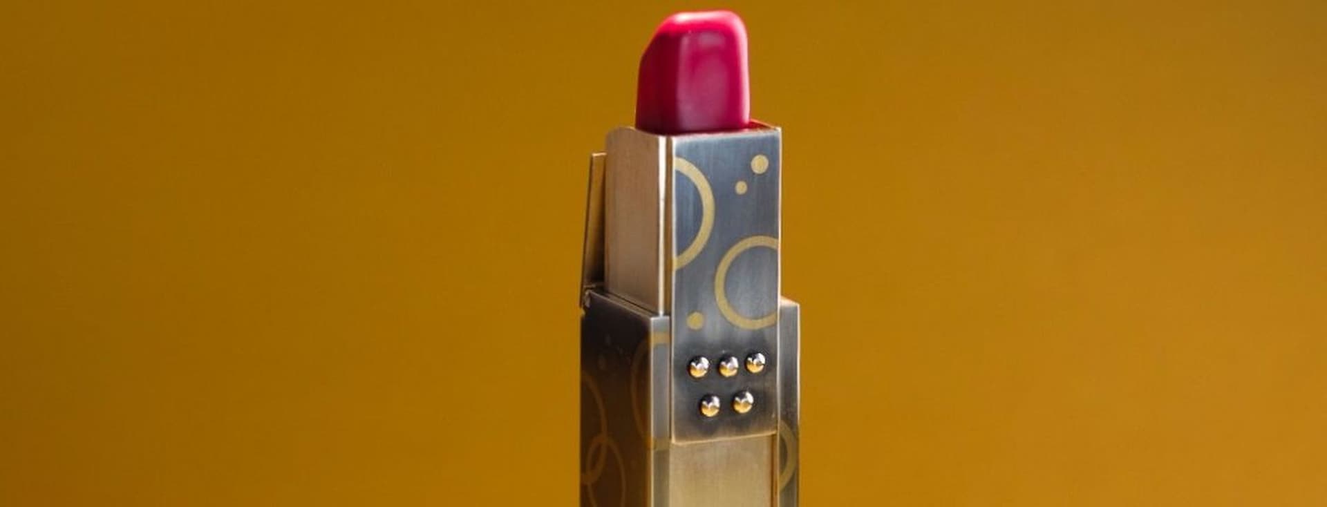 Roter Lippenstift mit Etui aus Gold und Silber mit geometrischem Muster vor goldbraunem Hintergrund.