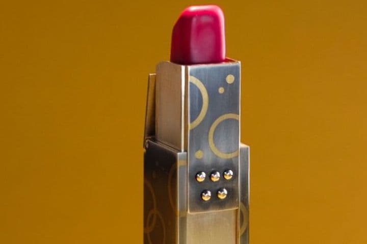 Roter Lippenstift mit Etui aus Gold und Silber mit geometrischem Muster vor goldbraunem Hintergrund.