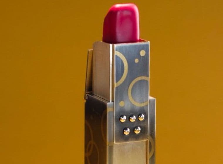 Roter Lippenstift mit Etui aus Gold und Silber mit geometrischem Muster vor goldbraunem Hintergrund.