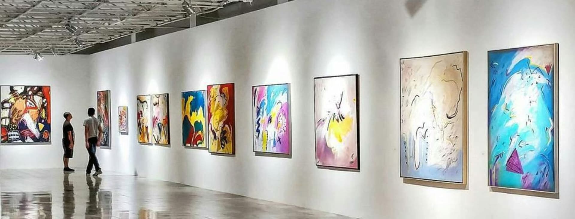 Kunstgalerie mit Besuchern, die farbenfrohe abstrakte Gemälde an weißen Wänden unter heller Beleuchtung betrachten.