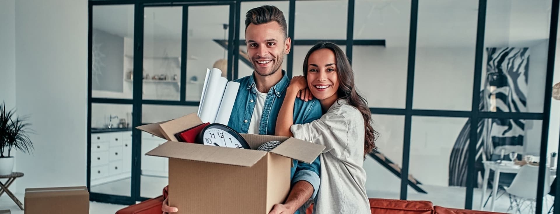 A smiling couple stands in a modern living room, holding a cardboard box filled with household items, including a clock and rolled papers.