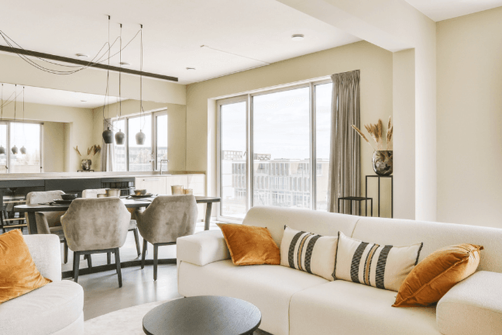 Bright, modern living room with a white sofa, orange pillows, dining table, and large windows.