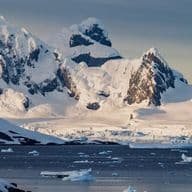 Snow-covered mountains and glaciers by a calm sea, with floating icebergs under a cloudy sky in a polar landscape.