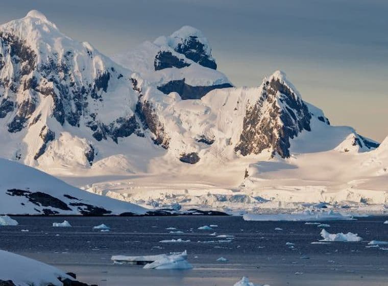 Snow-covered mountains and glaciers by a calm sea, with floating icebergs under a cloudy sky in a polar landscape.