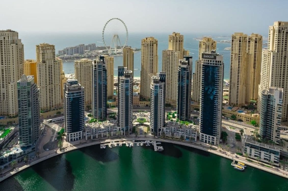 Aerial view of Dubai Marina's tall skyscrapers surrounding a curving waterfront, with a Ferris wheel visible in the background.