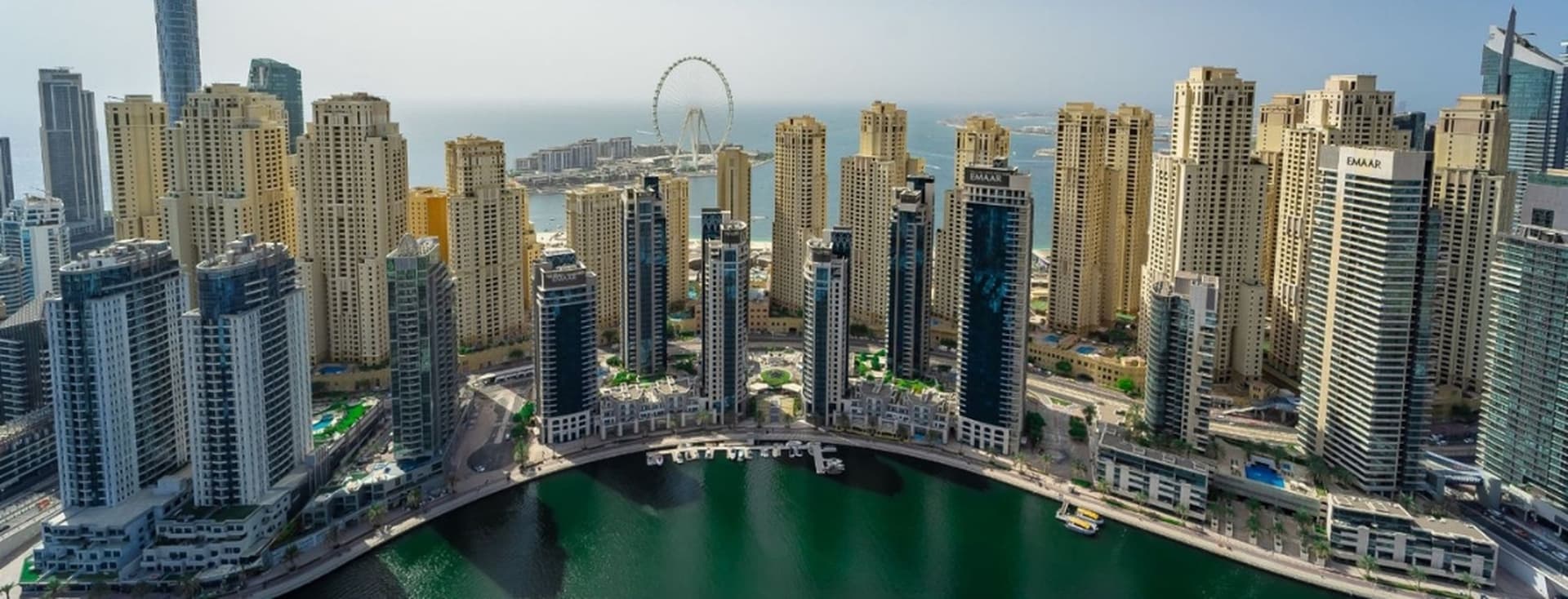 Skyline of tall, modern buildings surrounding a marina with a Ferris wheel in the background under a clear blue sky.