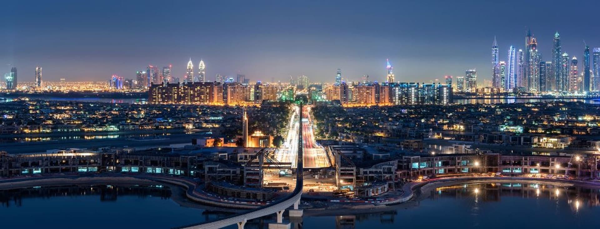 Panoramic view of Dubai at night, featuring illuminated skyscrapers, a lit bridge, and reflections on the water.