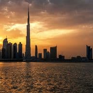 Ein dramatischer Sonnenuntergang über der Skyline Dubais, mit dem Burj Khalifa und umliegenden Wolkenkratzern als Silhouette vor goldenem Licht, das sich auf dem Wasser spiegelt.