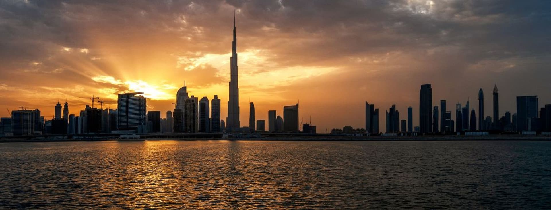 A dramatic sunset over the Dubai skyline, with the Burj Khalifa and surrounding skyscrapers silhouetted against golden light reflecting on the water.