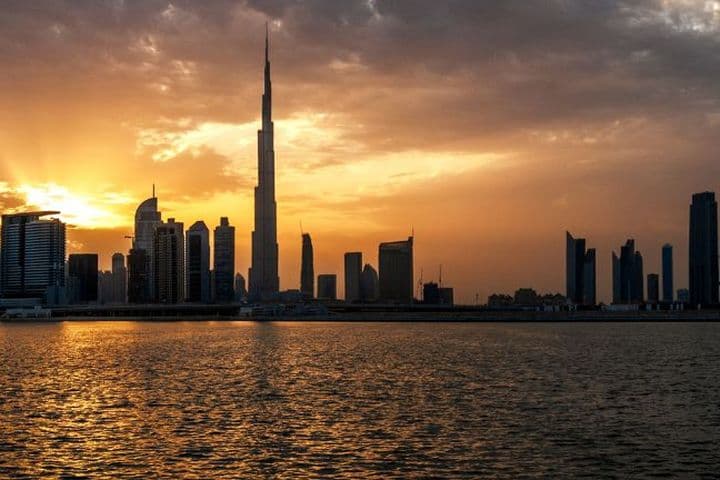 Ein dramatischer Sonnenuntergang über der Skyline Dubais, mit dem Burj Khalifa und umliegenden Wolkenkratzern als Silhouette vor goldenem Licht, das sich auf dem Wasser spiegelt.