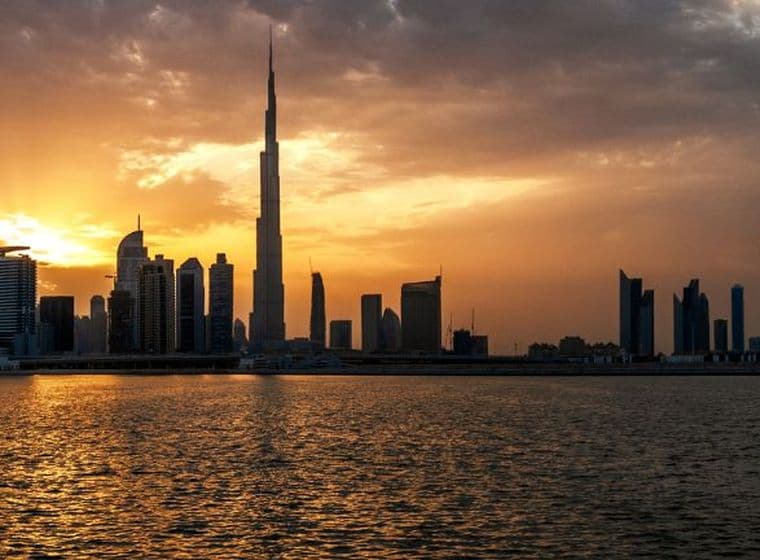 Ein dramatischer Sonnenuntergang über der Skyline Dubais, mit dem Burj Khalifa und umliegenden Wolkenkratzern als Silhouette vor goldenem Licht, das sich auf dem Wasser spiegelt.