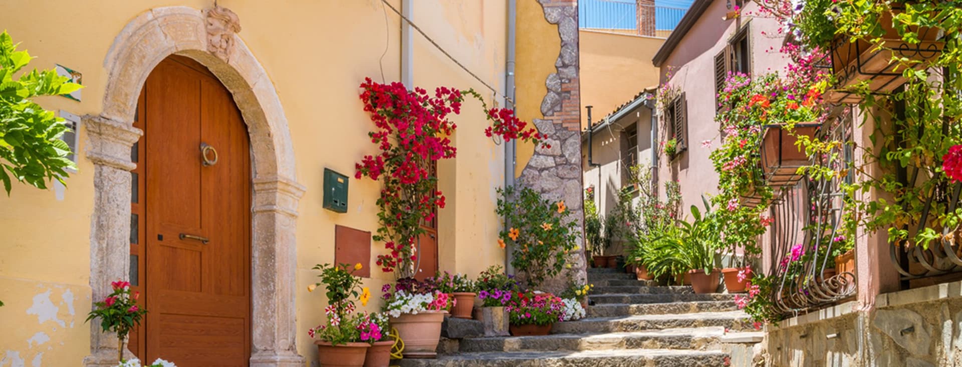 A charming narrow street with stone steps, vibrant flowers, colorful buildings with a wooden arch door, and lush vegetation.
