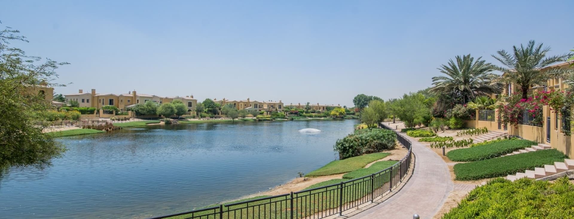A scenic view of a tranquil lake with a fountain, surrounded by lush greenery, pathways, and residential buildings under a clear blue sky.