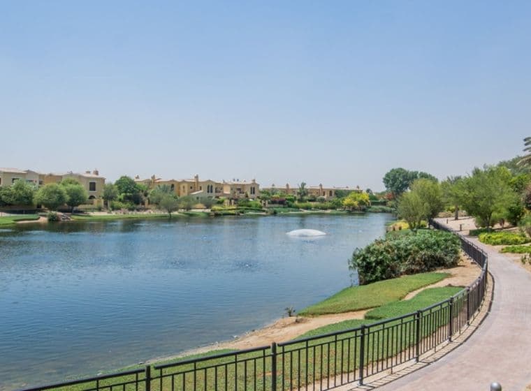 A scenic view of a tranquil lake with a fountain, surrounded by lush greenery, pathways, and residential buildings under a clear blue sky.