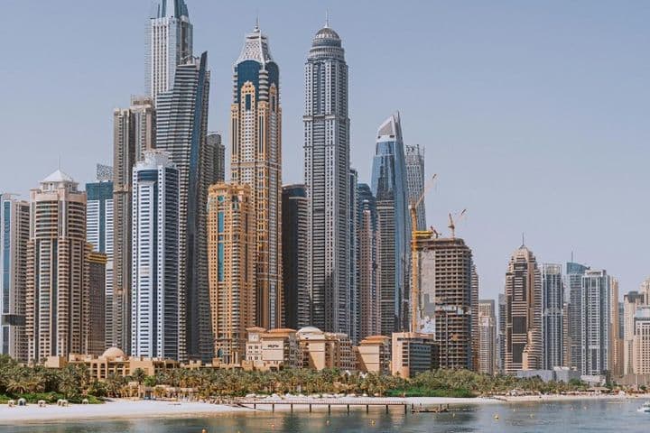 Skyline of a Dubai Marina housing market with tall skyscrapers, a waterfront, a beach, and clear blue skies.