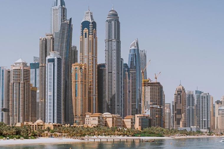 Skyline of a Dubai Marina housign market with tall skyscrapers, a waterfront, a beach, and clear blue skies.