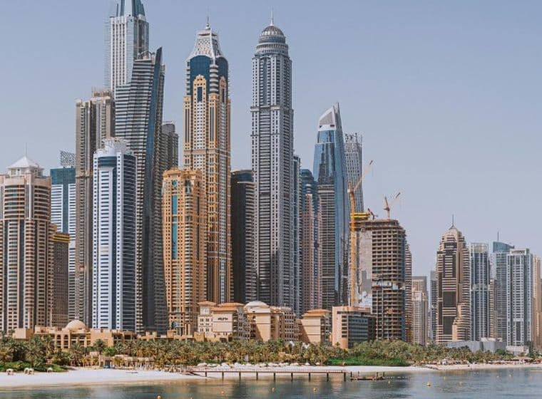 Skyline of a Dubai Marina housing market with tall skyscrapers, a waterfront, a beach, and clear blue skies.