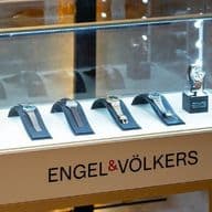 Display case with luxury watches on stands, illuminated under glass. The base is labeled "ENGEL & VÖLKERS."
