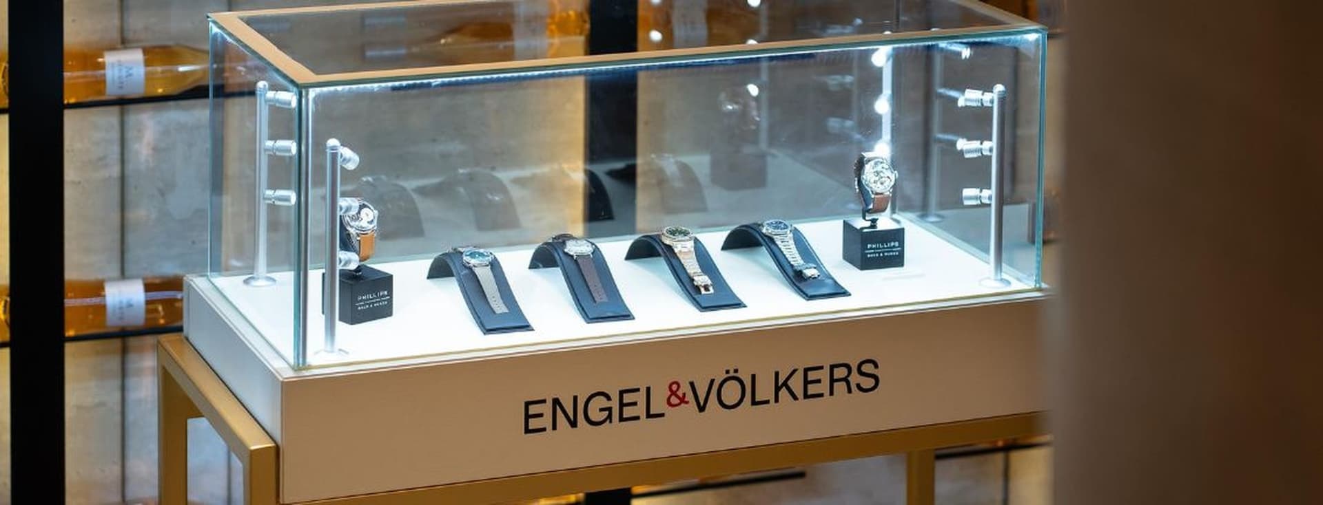 Display case with luxury watches on stands, illuminated under glass. The base is labeled "ENGEL & VÖLKERS."