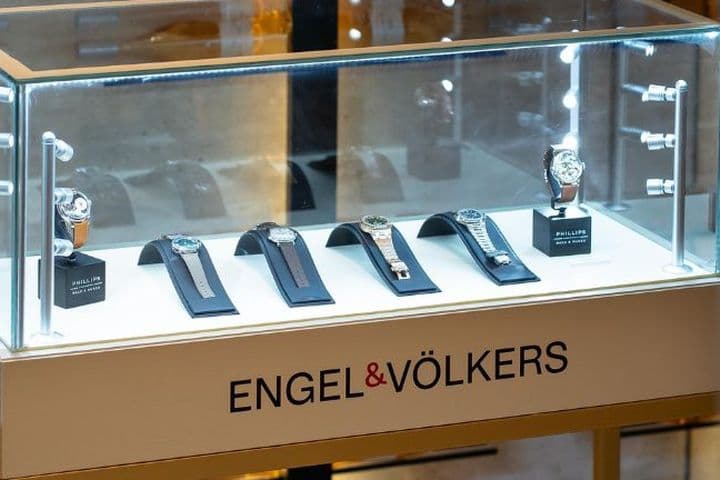 Display case with luxury watches on stands, illuminated under glass. The base is labeled "ENGEL & VÖLKERS."