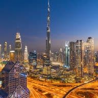 Aerial view of Downtown Dubai at dusk with illuminated skyscrapers and highways, featuring a prominent tall tower in the center.