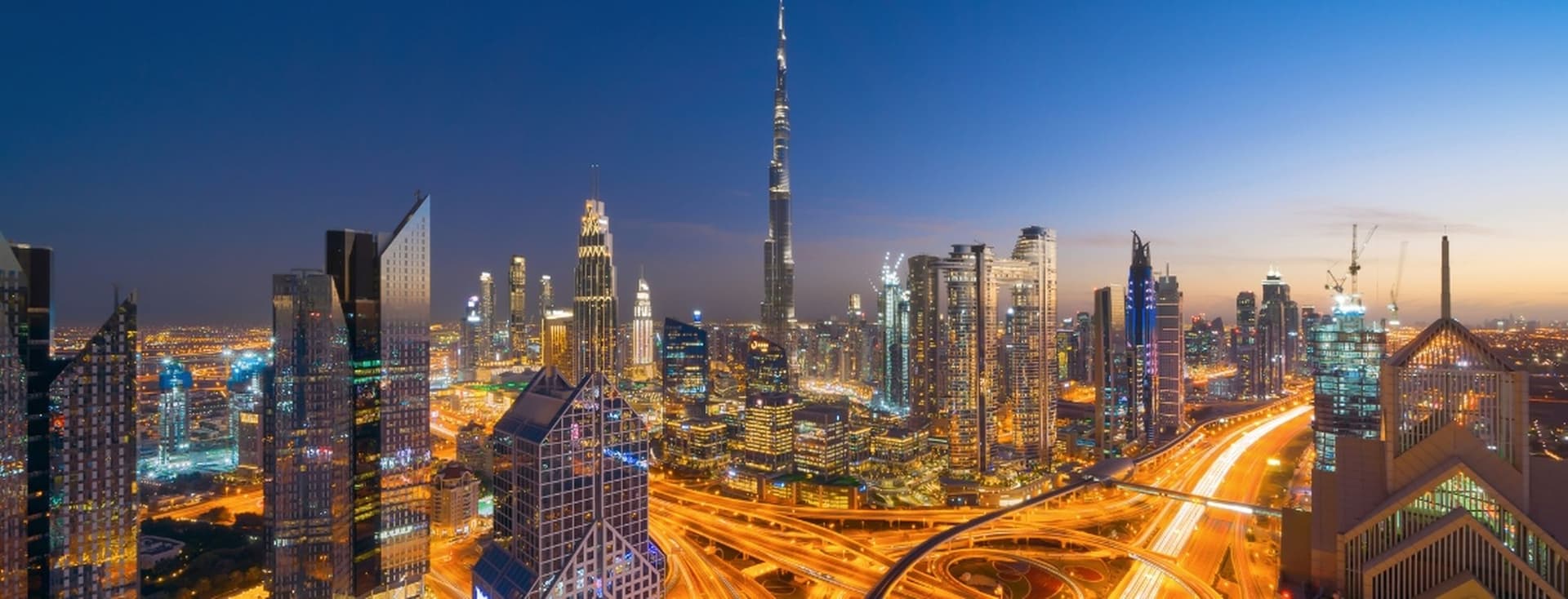 Aerial view of Downtown Dubai at dusk with illuminated skyscrapers and highways, featuring a prominent tall tower in the center.