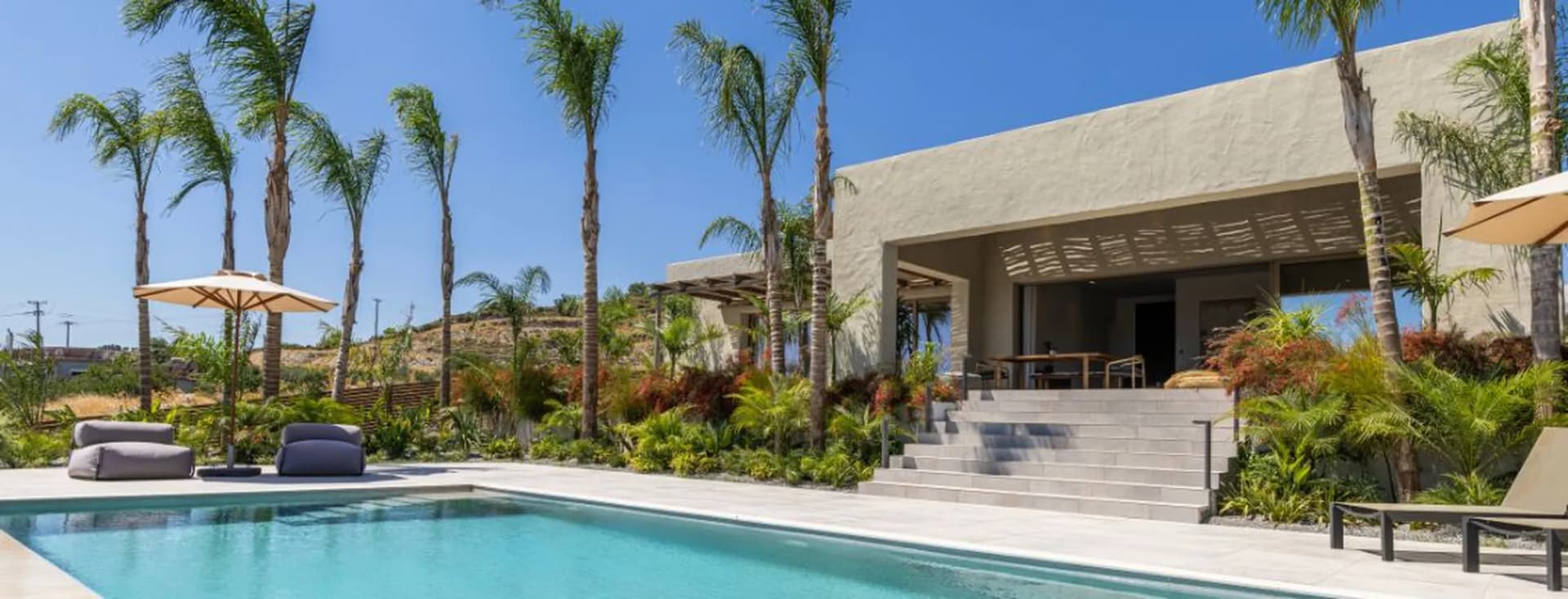 Modern luxury villa with a swimming pool, sun loungers, palm trees, and lush tropical landscaping under a clear blue sky.