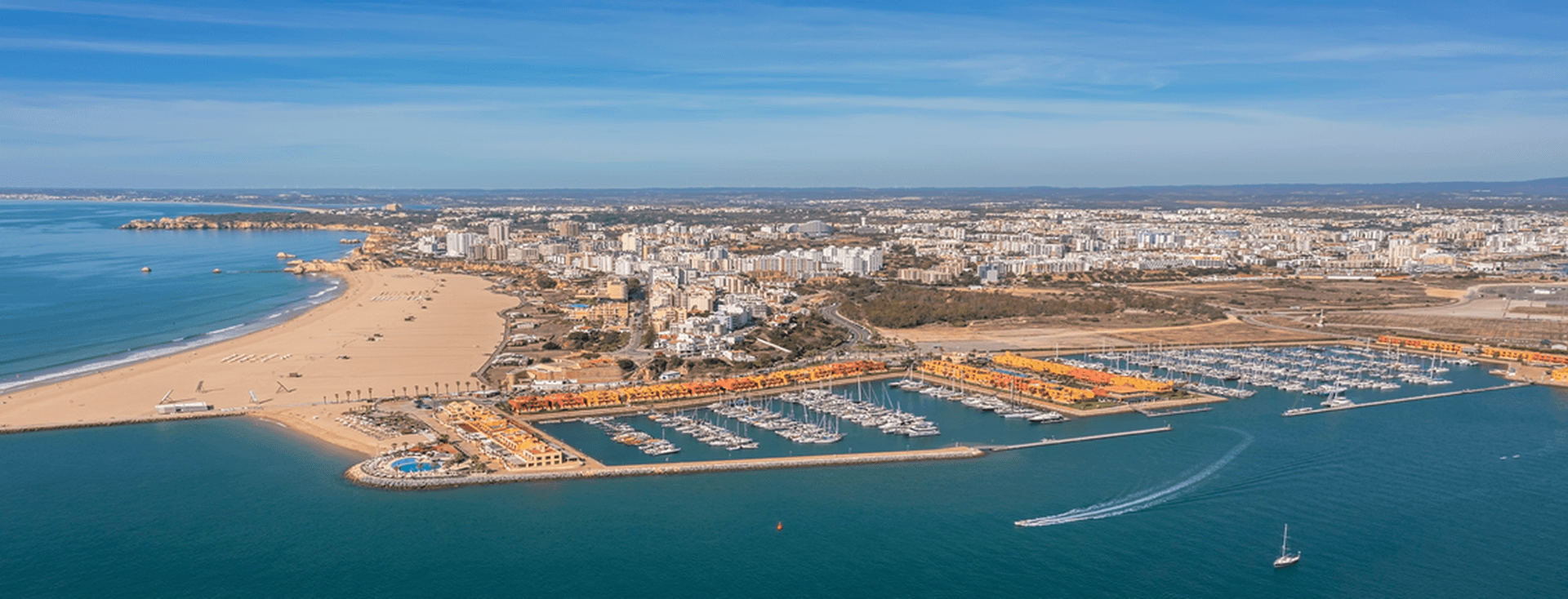 Luftaufnahme einer Küstenstadt mit einem Sandstrand, einem Yachthafen, bunten Gebäuden und Booten auf blauem Wasser unter einem klaren Himmel.