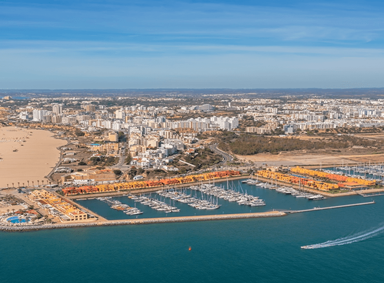 Luftaufnahme einer Küstenstadt mit einem Sandstrand, einem Yachthafen, bunten Gebäuden und Booten auf blauem Wasser unter einem klaren Himmel.