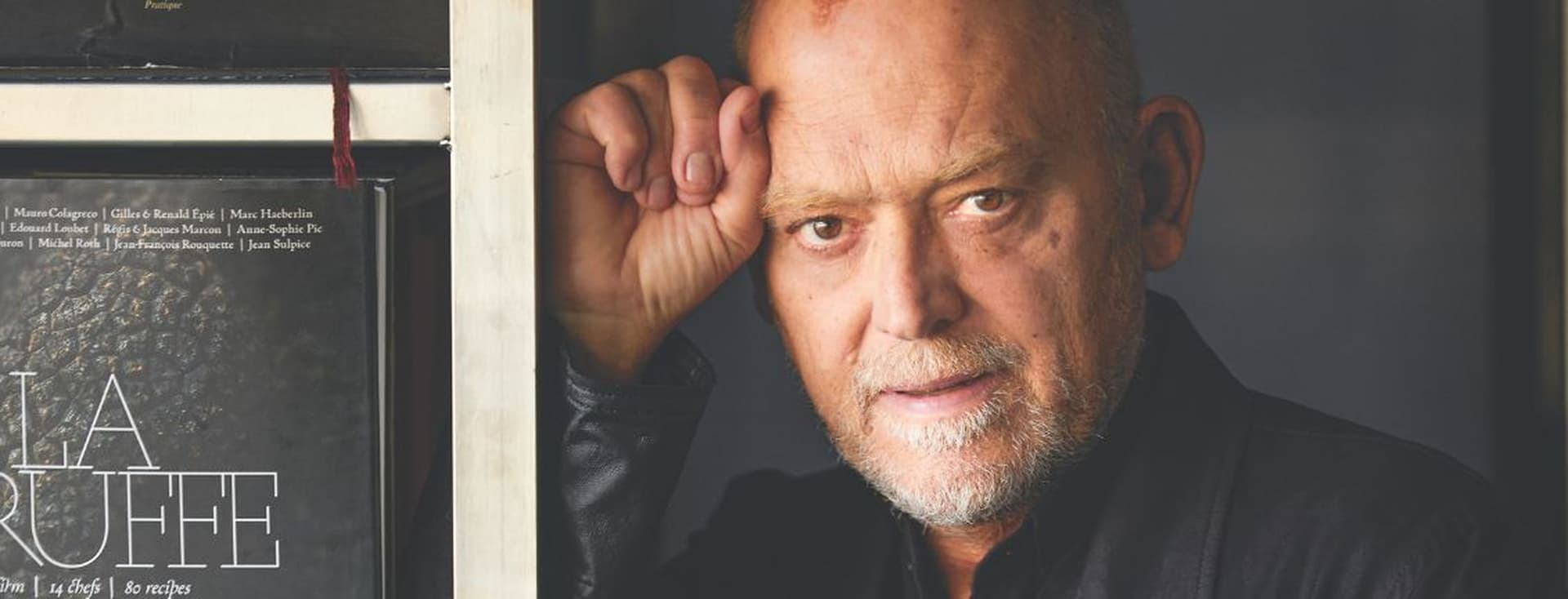 Portrait of a middle-aged man with a thoughtful expression, leaning against a bookshelf next to a cookbook, in a softly lit interior.