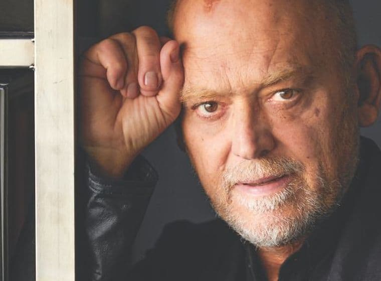 Portrait of a middle-aged man with a thoughtful expression, leaning against a bookshelf next to a cookbook, in a softly lit interior.
