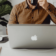 A person in a plaid shirt drinking from a mug at a white desk with a laptop and office supplies.