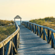 Un pontó de fusta és l'entrada a la platja, amb vegetació salvatge a banda i banda.