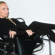 Γυναίκα με μακριά ξανθή κοτσίδα, φοράει εφαρμοστό μαύρο σύνολο και γόβες με μυτερή μύτη, είναι ξαπλωμένη σε μοντέρνα μαύρη μεταλλική καρέκλα και κρατά δομημένη μαύρη τσάντα, με λευκό φόντο.