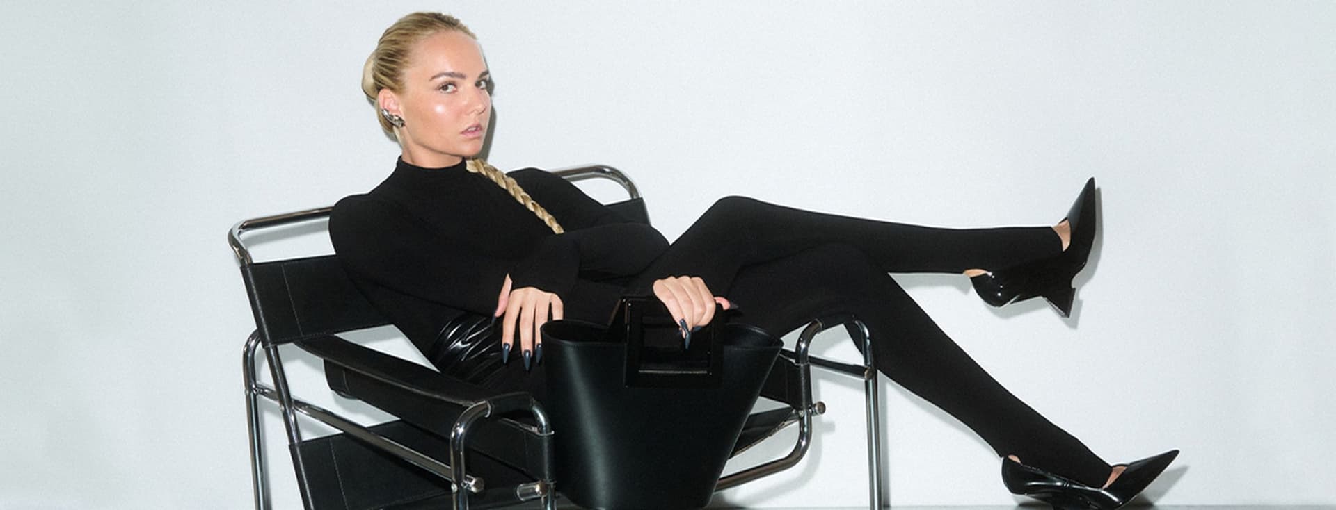 Γυναίκα με μακριά ξανθή κοτσίδα, φοράει εφαρμοστό μαύρο σύνολο και γόβες με μυτερή μύτη, είναι ξαπλωμένη σε μοντέρνα μαύρη μεταλλική καρέκλα και κρατά δομημένη μαύρη τσάντα, με λευκό φόντο.